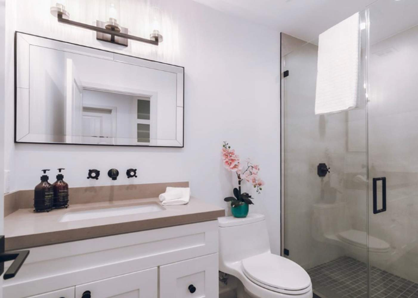 A modern bathroom with a white vanity, large mirror, glass-enclosed shower, and decorative orchid, showcasing Smith & Son Construction's work in Martinez.