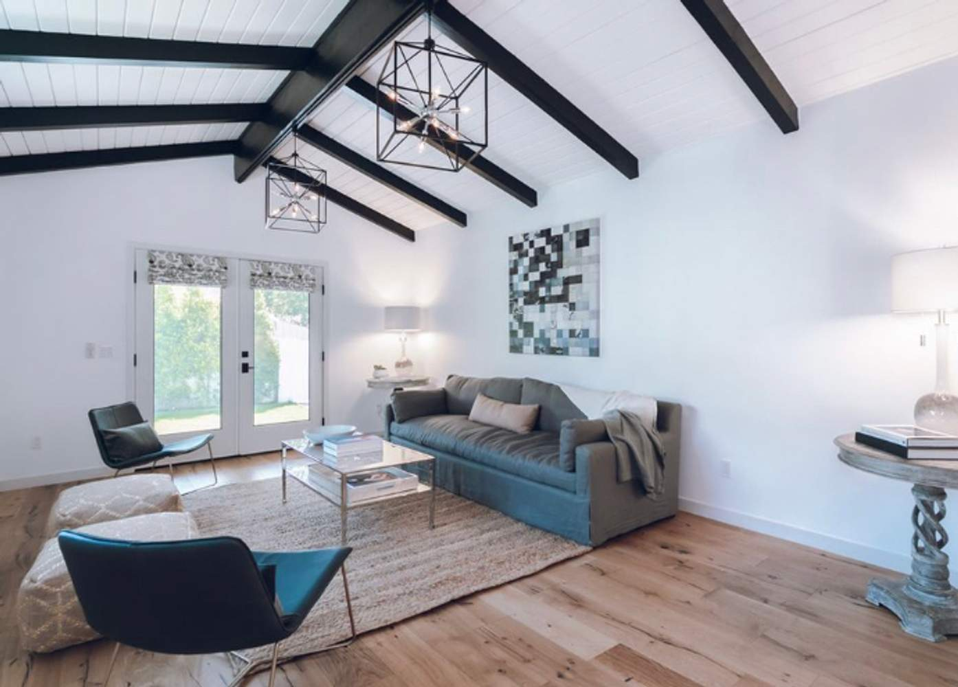 A modern living room with a vaulted ceiling featuring exposed wooden beams, furnished with a gray sofa, two chairs, a lamp, and contemporary art.