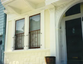 A well-maintained house facade with intricate wooden and stone detailing, featuring two windows with decorative iron grilles.