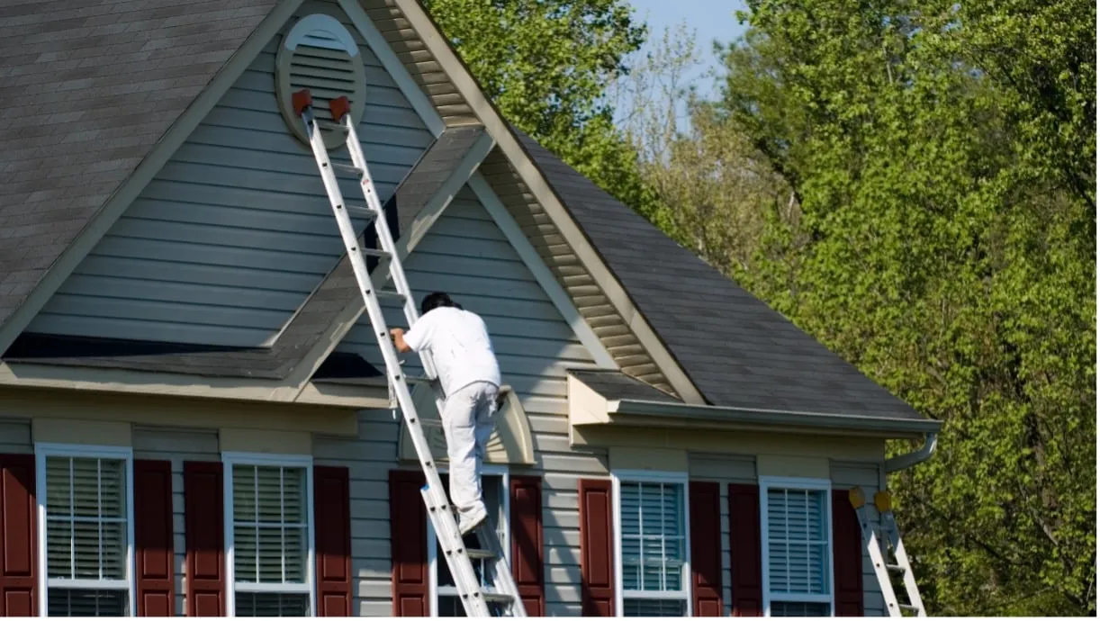 A painter from Stanford Painting, Inc. works on a house exterior in Mountain View, standing on a ladder and painting near the eaves.