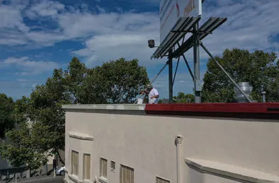 A person standing on the roof of a building, near a large billboard, surrounded by trees and a partly cloudy sky.