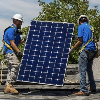 Two workers in safety gear installing a solar panel on a rooftop, surrounded by trees, representing sustainable energy efforts by SunMade Smart Roofing, Solar & Electric in Fresno.