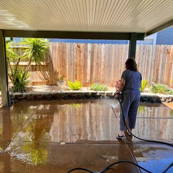 A person washing a patio under a covered structure with sunlight reflecting on the wet surface, featuring a garden area in the background.