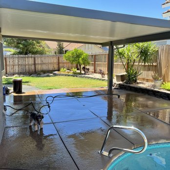A patio with a covered awning next to a small pool, surrounded by a well-kept garden and a wooden fence, under a clear blue sky.