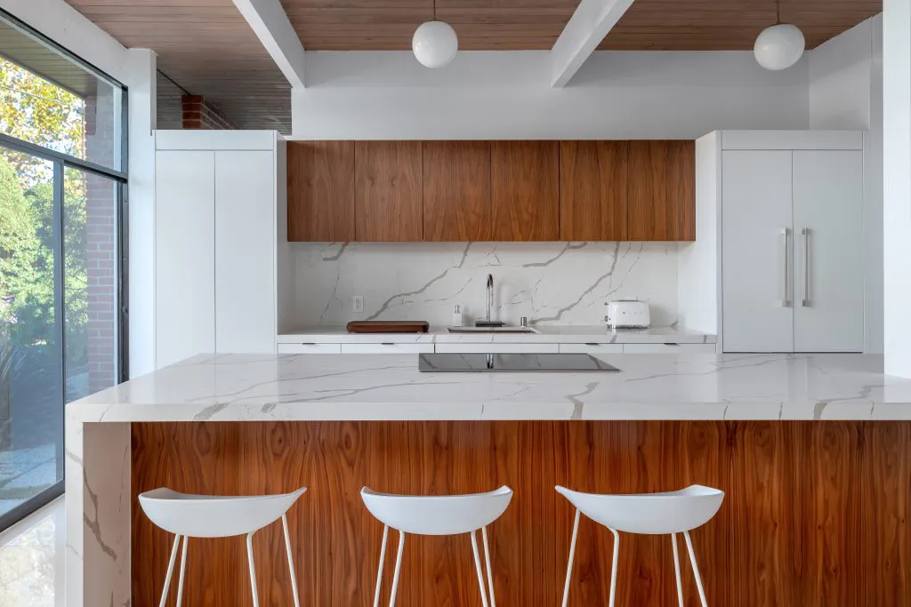 Modern kitchen with white counters and wooden cabinets, featuring pendant lighting and three white stools at the island.