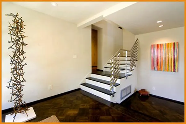 A modern interior with a staircase featuring metal railings, a vertical abstract sculpture on the floor, and a colorful abstract painting on the wall.