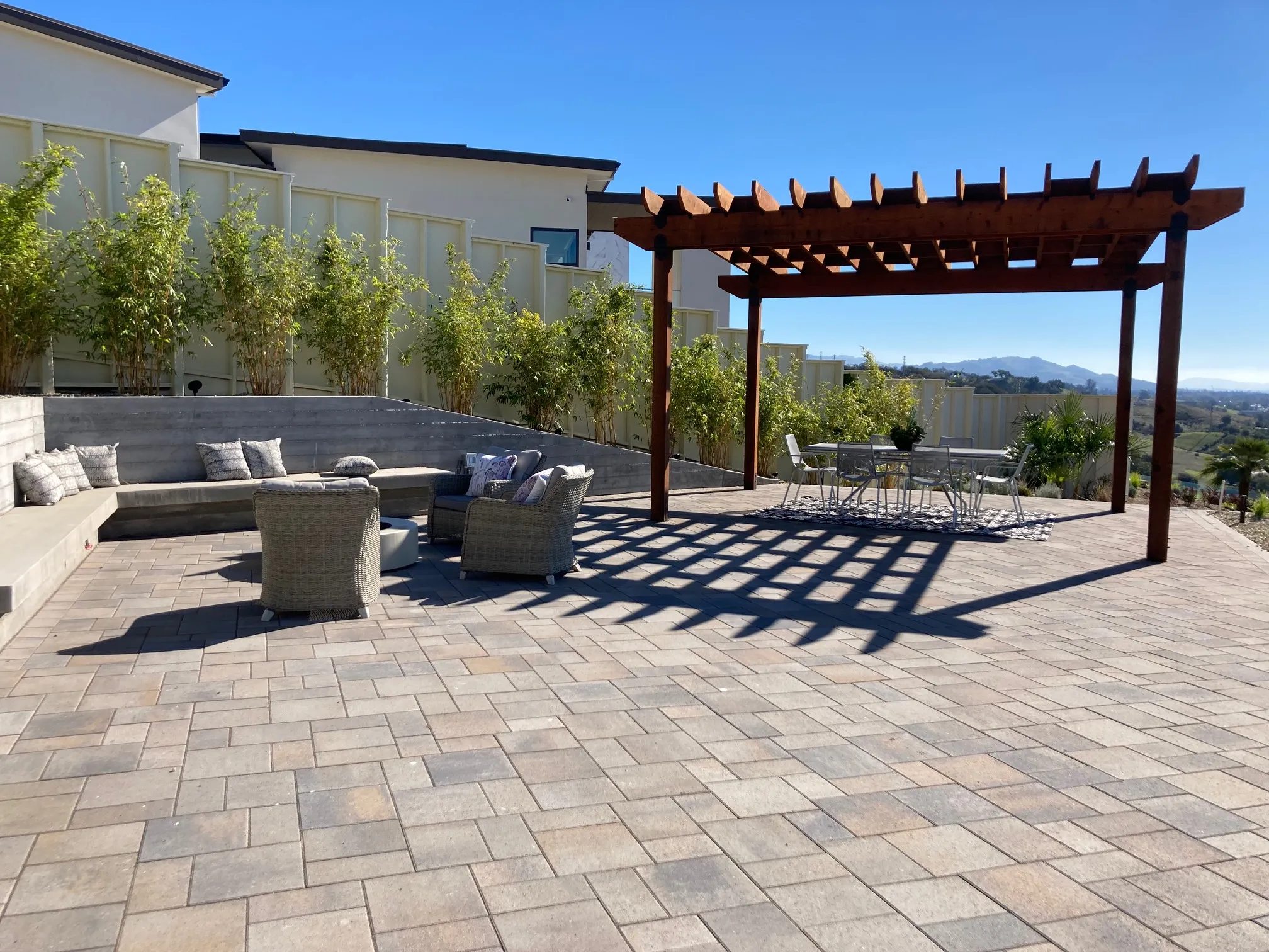 A patio with wicker chairs and a table under a wooden pergola, surrounded by lush greenery, designed by The Legacy Paver Group in Santa Rosa.
