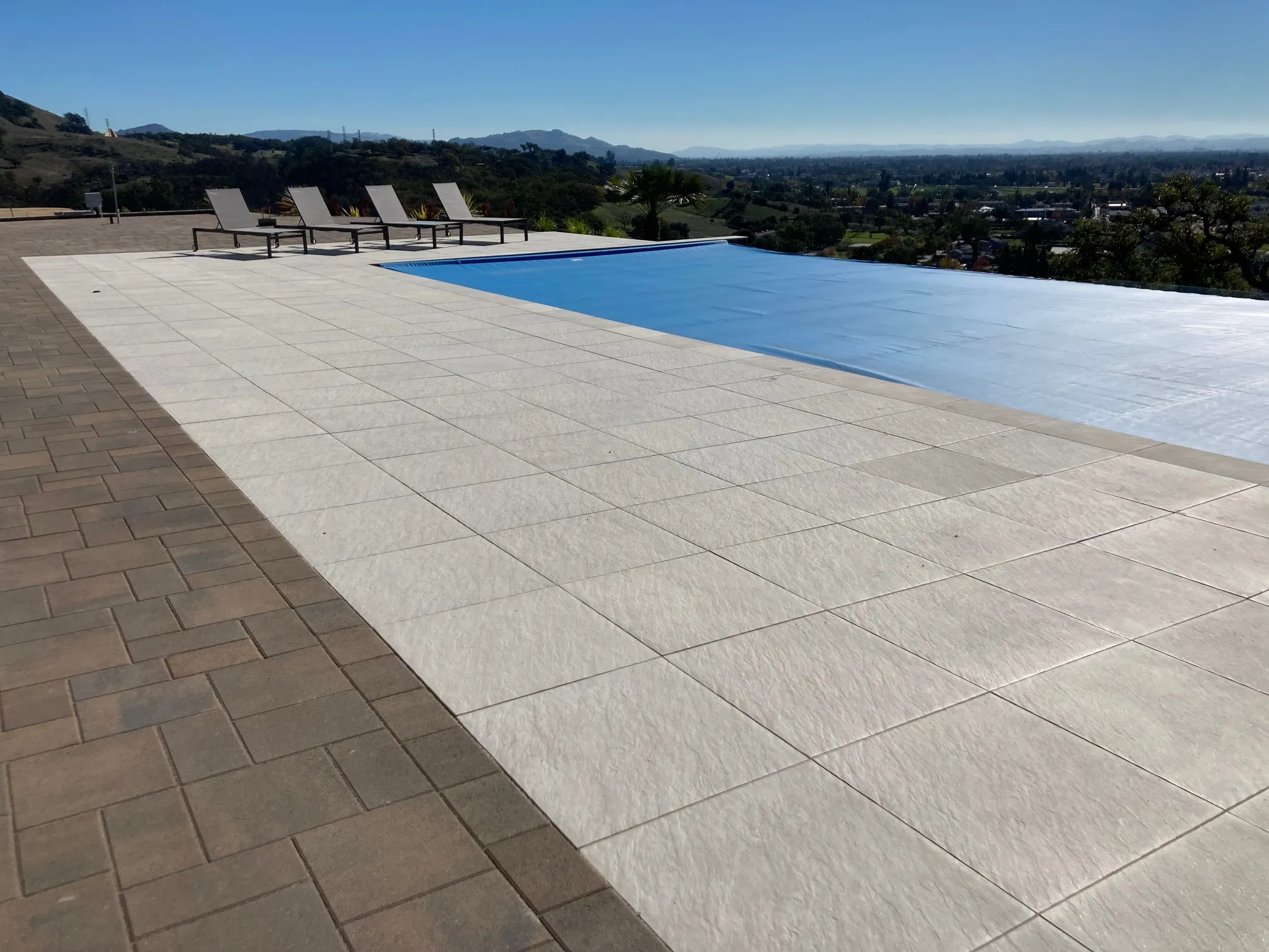 A modern patio area by a serene pool, featuring pavers installed by The Legacy Paver Group in Santa Rosa, with distant hills and a clear sky in the background.