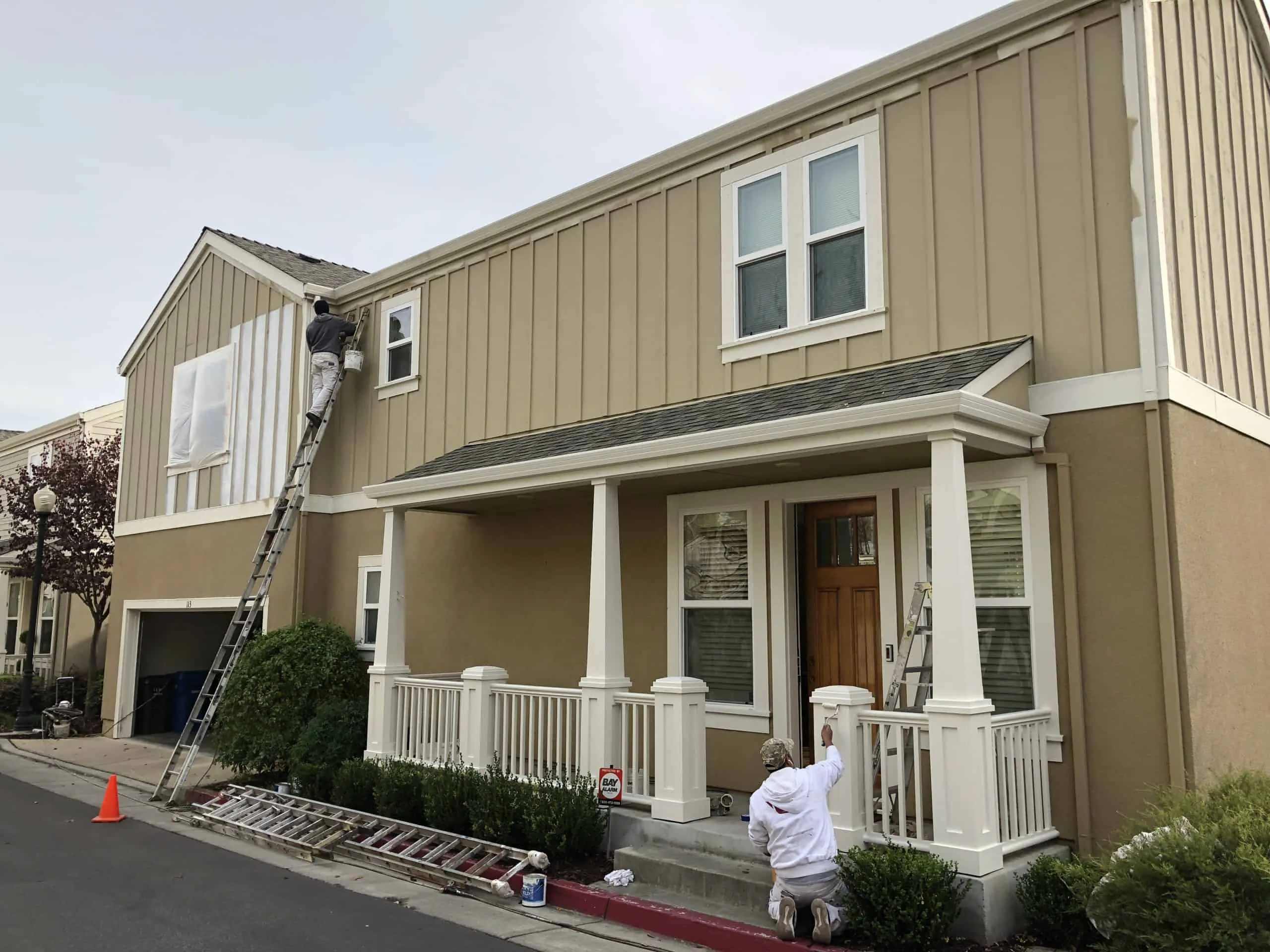 Two painters from The Painting Pros work on a house exterior in Los Gatos; one on a ladder painting the upper wall, and another kneeling and painting the porch.