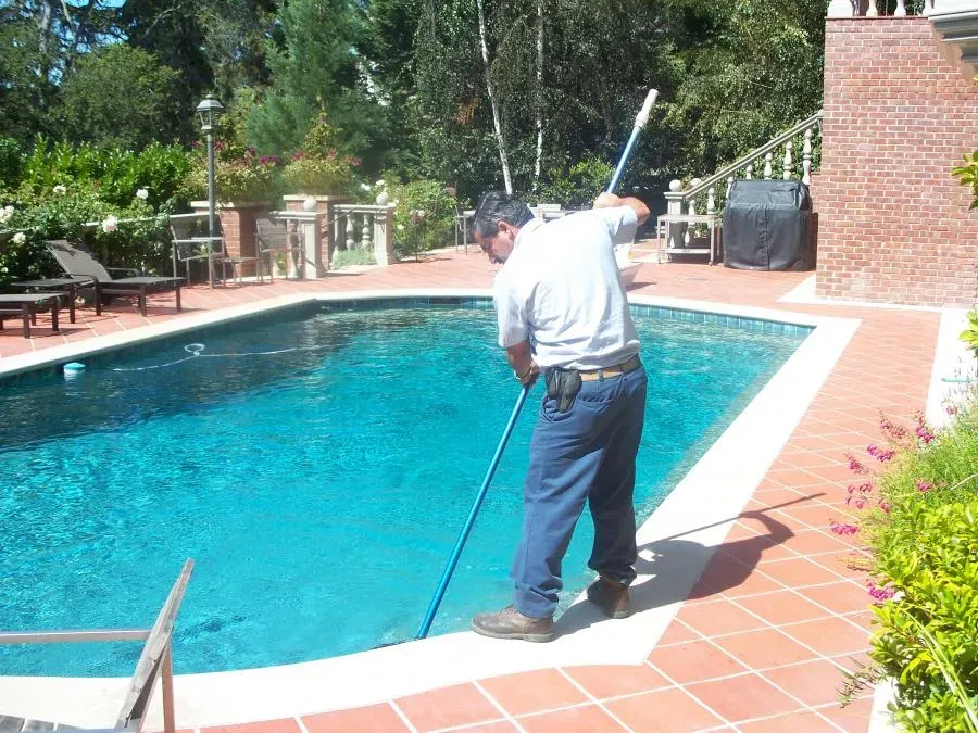 "A professional from The Pool Doctor cleans a residential pool with a long tool in El Sobrante, surrounded by greenery."