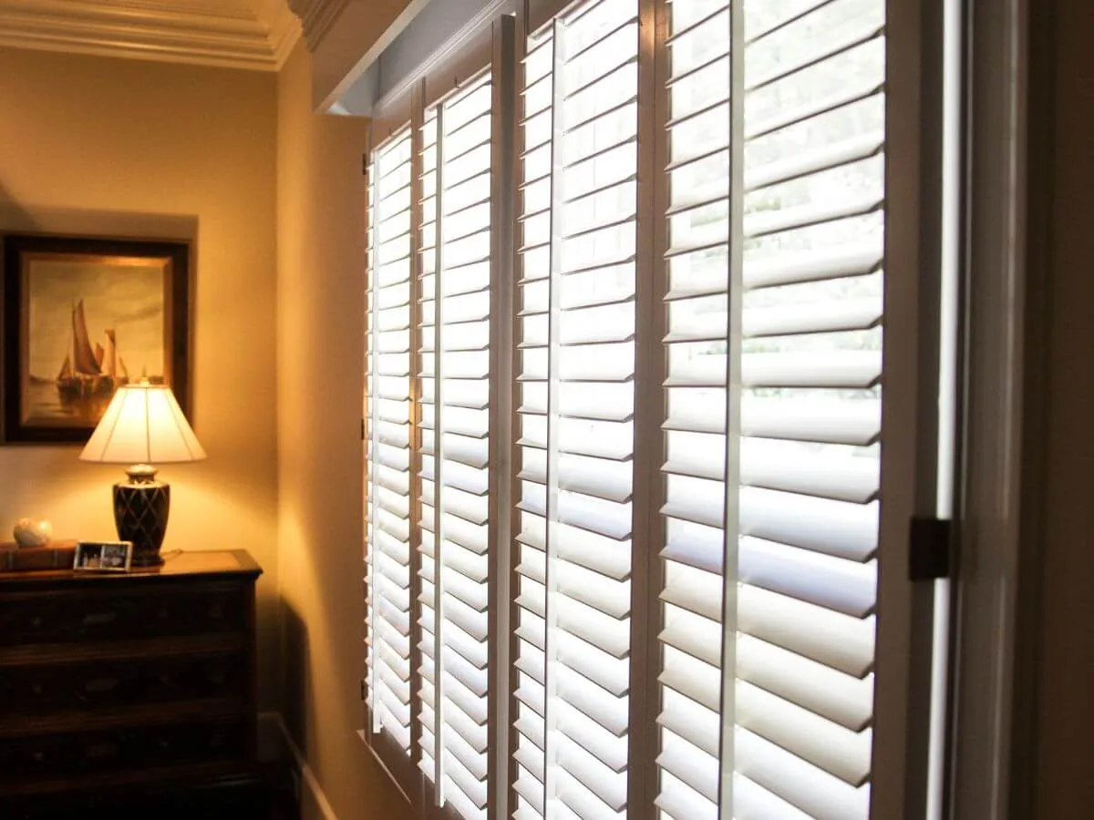 Room interior with white window shutters partially open, designed by The Window Design Studio in Petaluma, illuminating a lamp and framed painting.