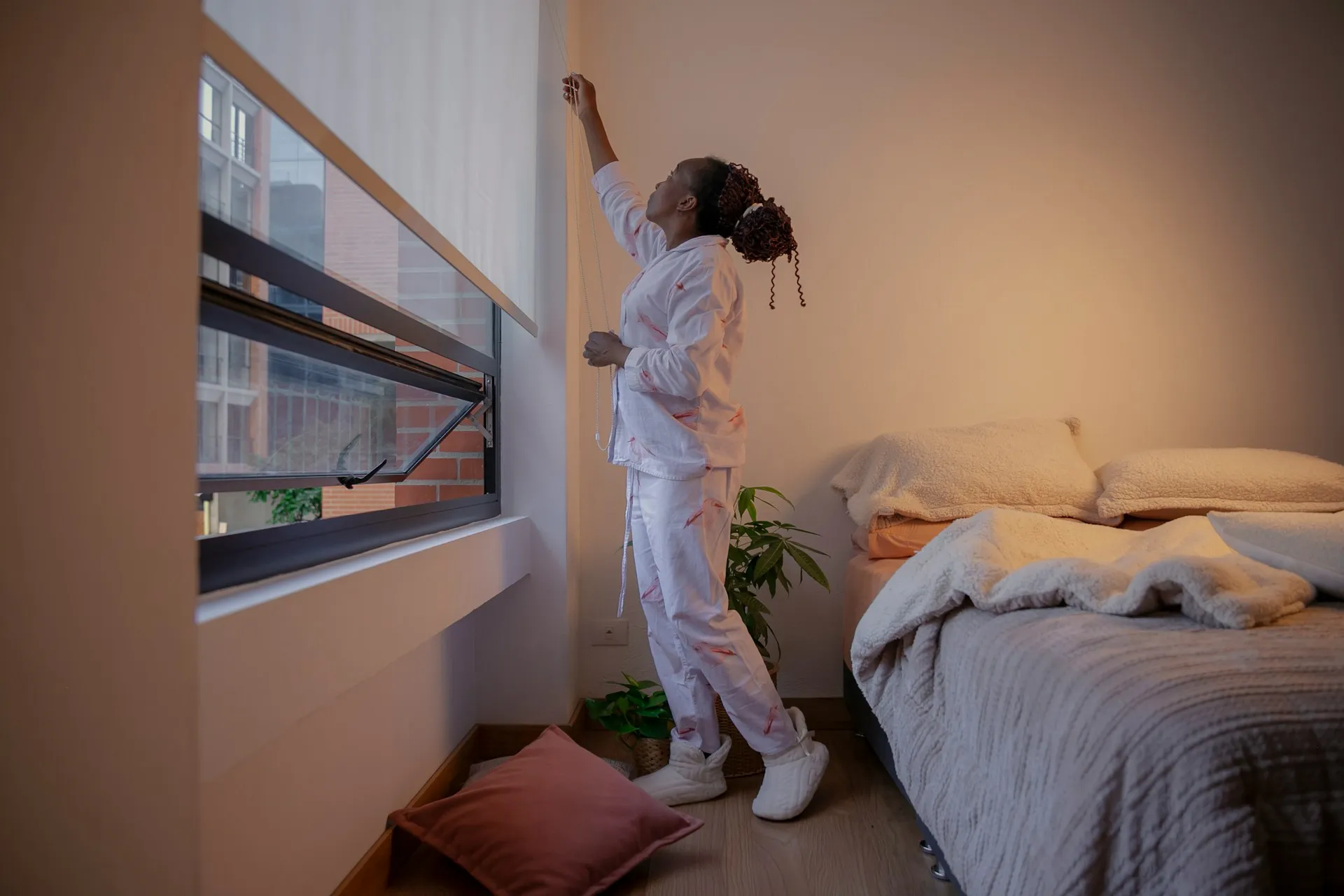 A person in pajamas adjusts a window blind in a cozy bedroom setting, featuring a neatly made bed and a modern-looking window design.