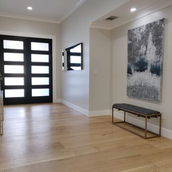 A modern foyer with wooden flooring, a black multi-panel door, abstract art on the wall, and a bench, showcasing the interior design by True Builders in San Jose.
