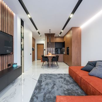 A modern living room and kitchen area with wooden accents, featuring a gray rug, red sofa, and a sleek dining set, designed by True Builders in San Jose.