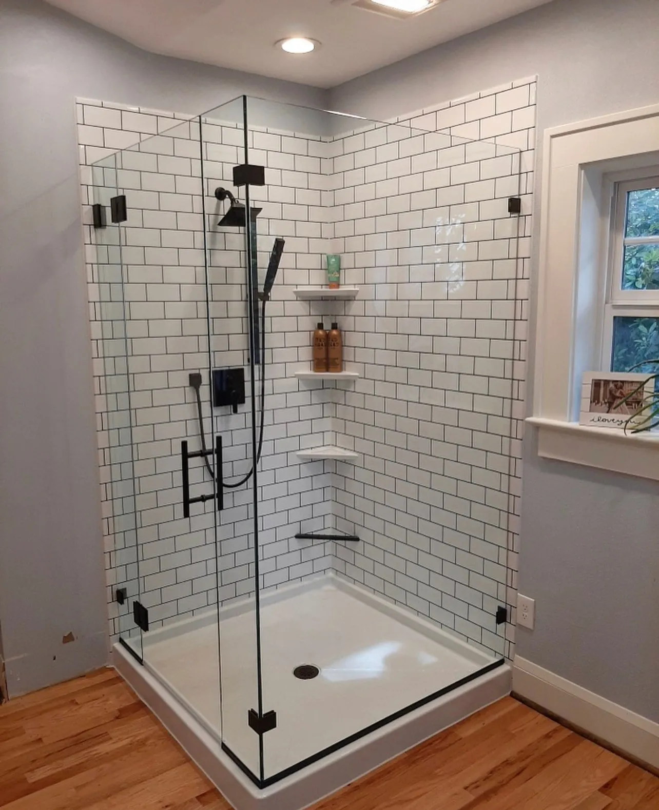 A modern glass-enclosed shower featuring white subway tiles, a black showerhead, and neatly arranged shelves in a bathroom setting.