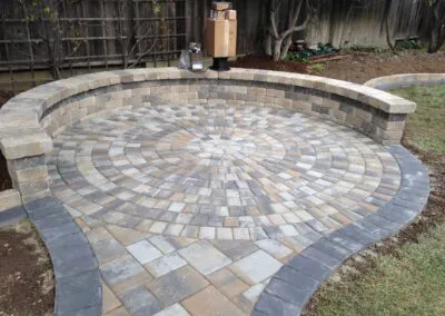 A circular patterned patio with neatly arranged pavers and a curved sitting wall, designed by Unique Pavers in a garden setting.