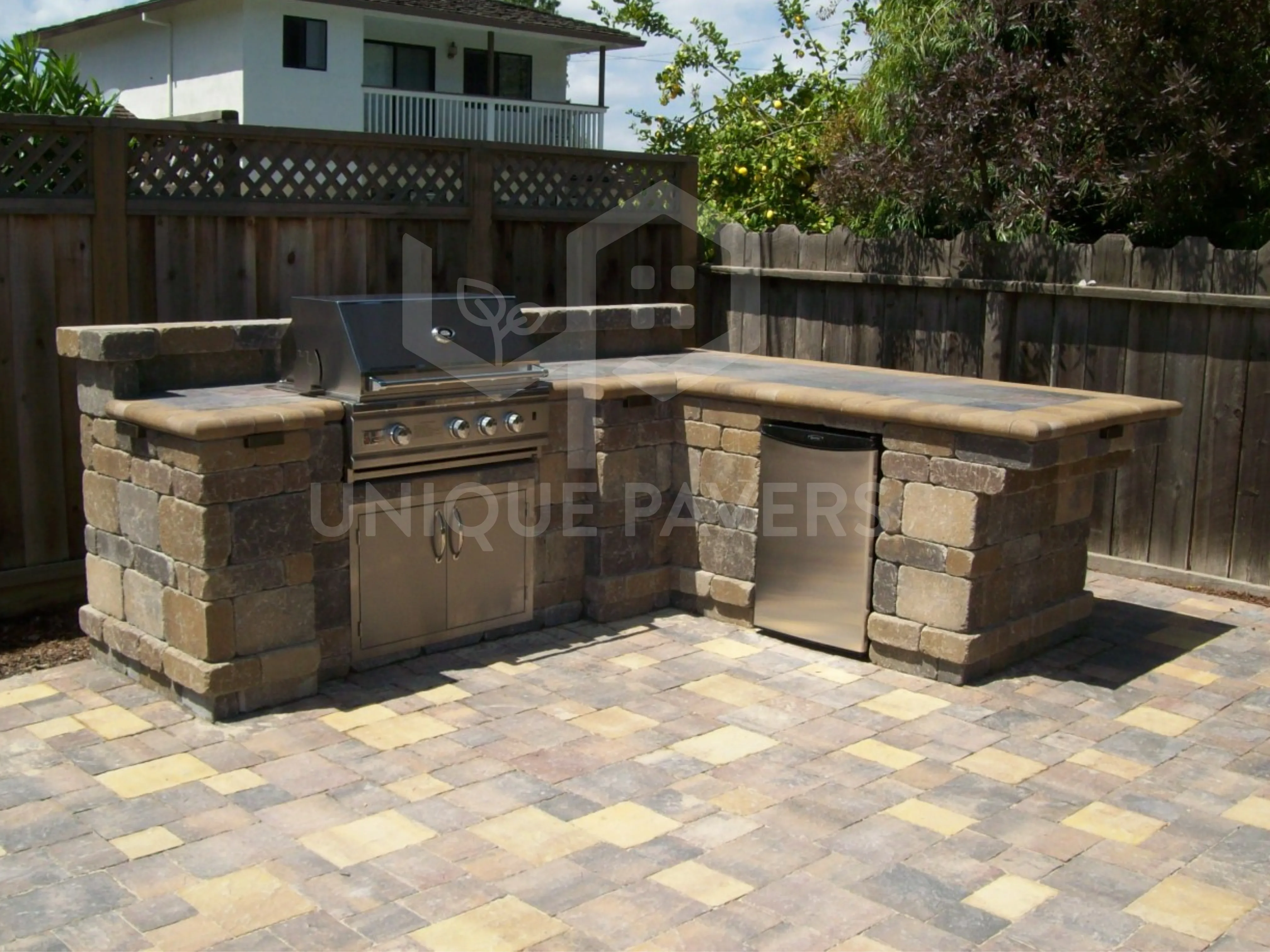 Outdoor stone kitchen in a San Jose backyard, featuring a stainless steel grill and fridge, with stone countertops and paver flooring, crafted by Unique Pavers.