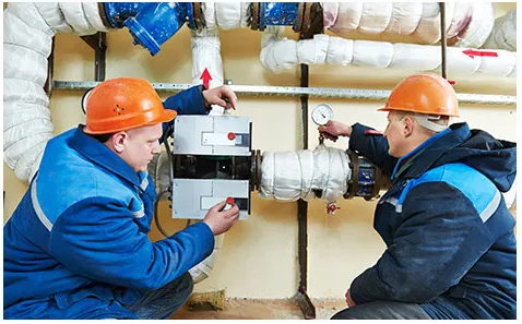 Two technicians in orange helmets and blue uniforms inspecting and adjusting HVAC equipment wrapped in insulation.
