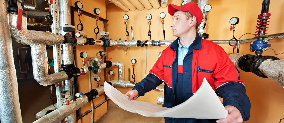 Technician in a red cap and jacket examining HVAC piping and gauges, holding a blueprint, in a mechanical room.