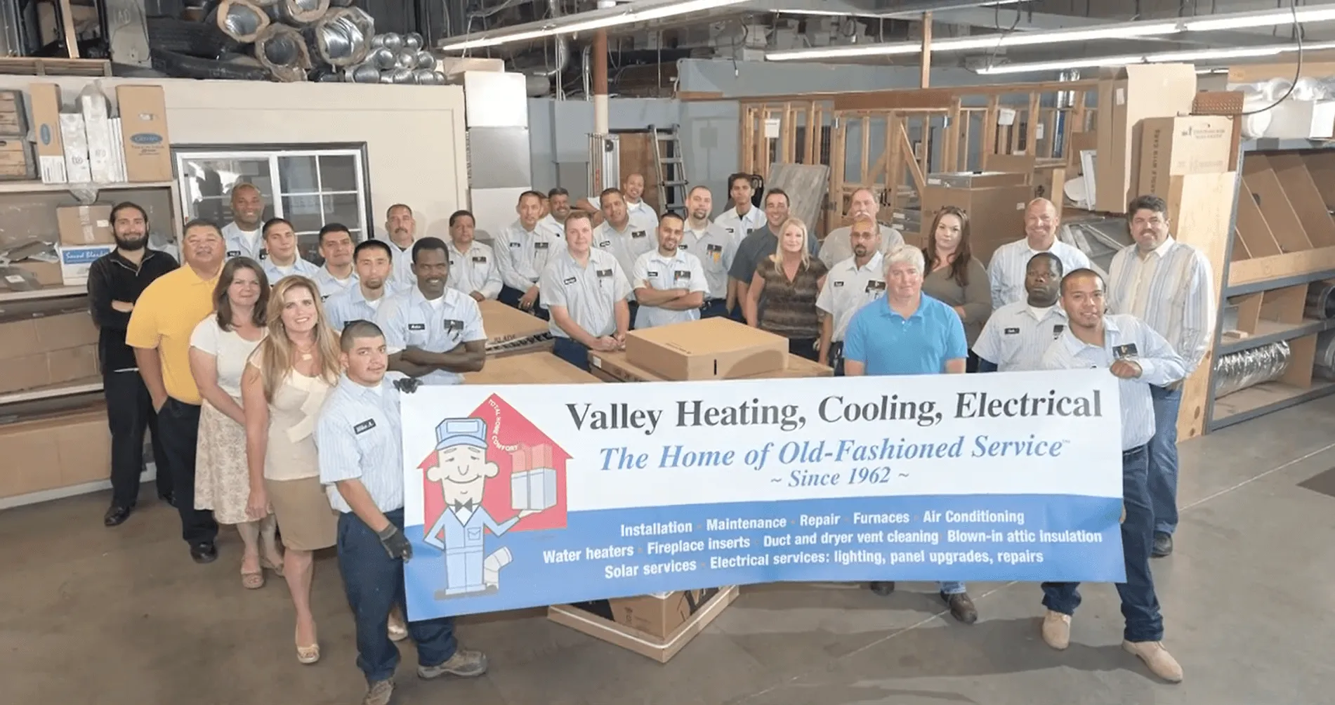 A group of employees from Valley Heating, Cooling, Electrical and Solar in San Jose stand together in a workshop, holding a banner showcasing their services.