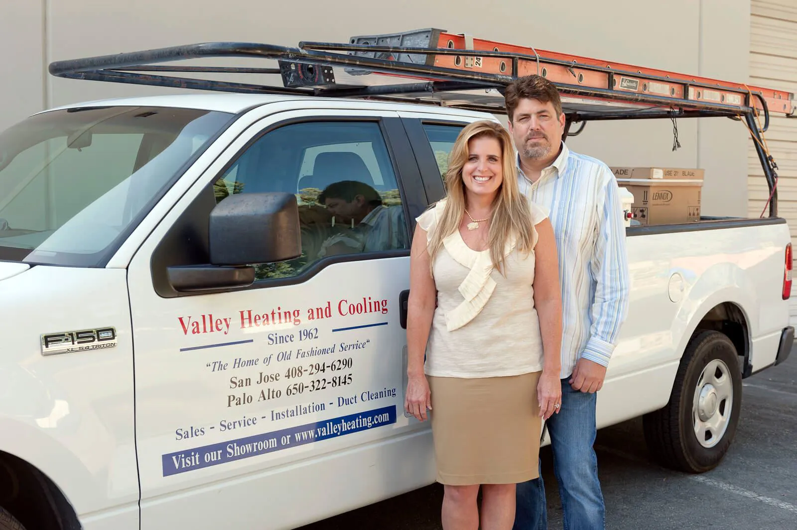 A man and woman stand smiling in front of a Valley Heating, Cooling, Electrical and Solar service vehicle with a ladder on top, representing their commitment to service in San Jose.