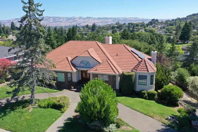 A red-roofed home surrounded by greenery, showcasing WedgeRoofing's craftsmanship in Petaluma. The house is set against a backdrop of hills and trees.