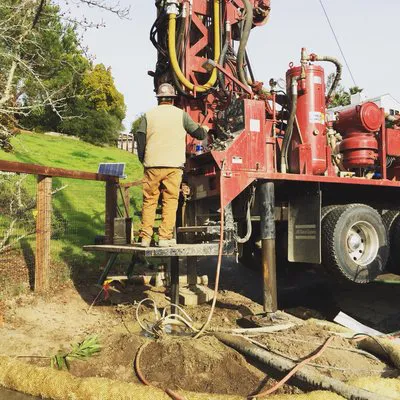 A worker from Weeks Drilling & Pump Company operates a drilling rig in a grassy area in Sebastopol, with equipment and hoses visible on the ground.