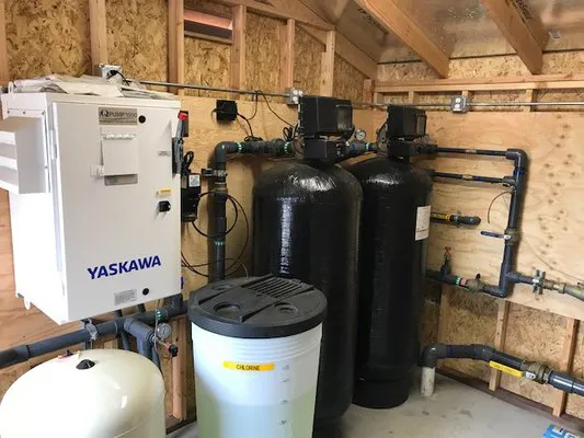 An indoor setup of a water filtration and pump system, featuring large black tanks, a Yaskawa control unit, and various pipes, installed by Weeks Drilling & Pump Company.