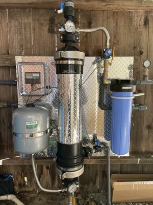 An advanced water filtration and pump system installed on a wooden wall, featuring a metal pressure tank labeled "Weeks Drilling & Pump Company" and a blue filter unit.