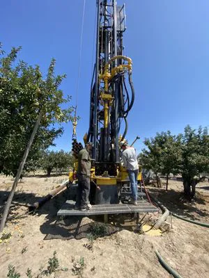 Two workers operate a large drilling rig in a field, surrounded by trees, under a clear blue sky, representing Weeks Drilling & Pump Company's operations in Sebastopol.