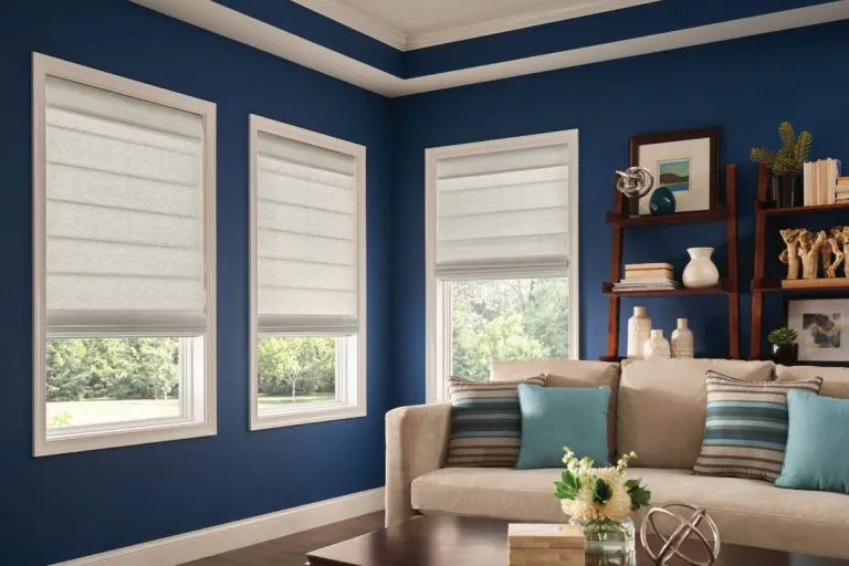 A living room with blue walls featuring three windows with white Roman shades, a beige couch with striped and solid pillows, and a wooden shelf holding books and decor items.