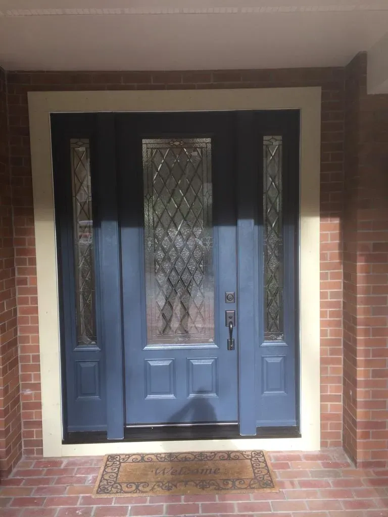 A blue front door with decorative glass panels and a welcome mat, surrounded by brick walls.