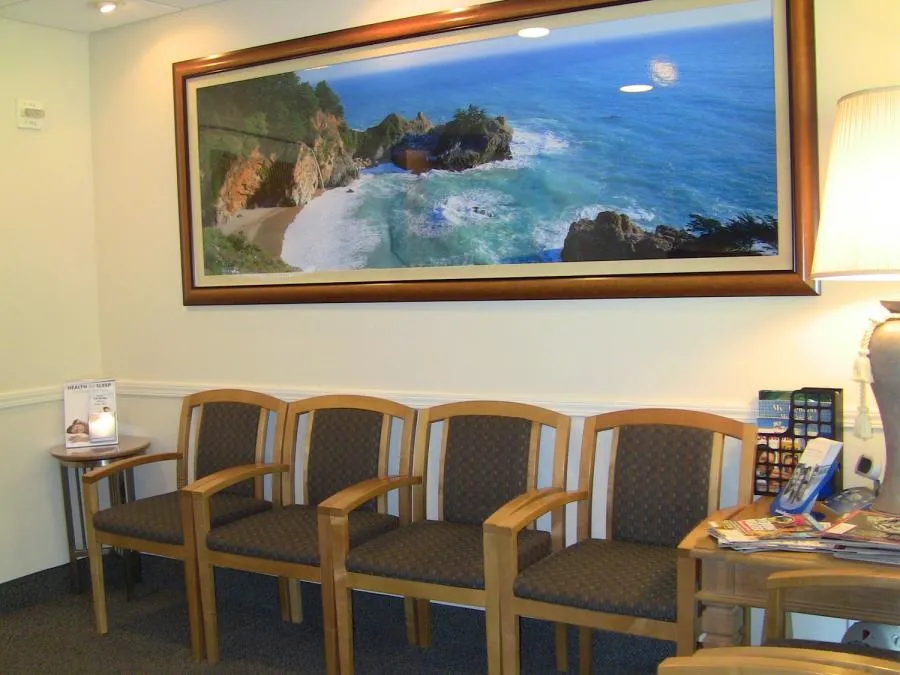 A waiting room in the William M. Schneider, D.D.S. office in Walnut Creek, featuring a row of wooden chairs, a side table with magazines, and a large framed coastal photo on the wall.