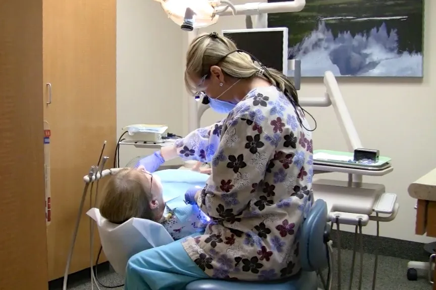 A dental professional in patterned scrubs attends to a patient in a dental chair, under a bright examination light.
