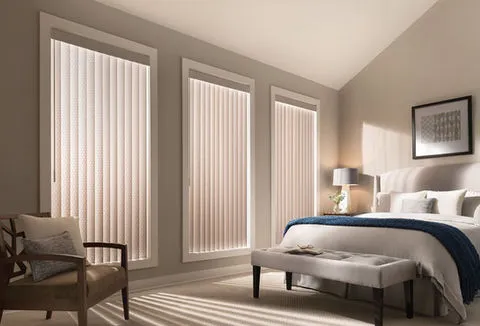 A bedroom with vertical blinds on three large windows, featuring a neatly made bed with a blue blanket, a bench at the foot, a lamp, and a chair.