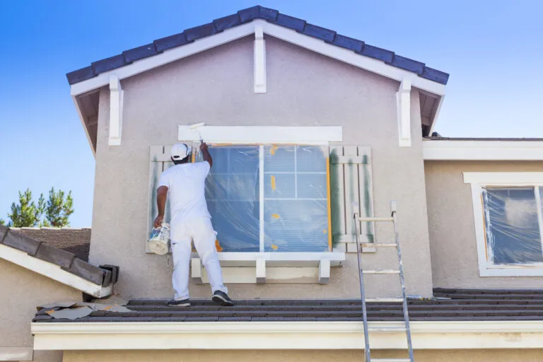 A painter from Wine Country Painters, Inc. applies a fresh coat of paint to the exterior window frame of a house in Sebastopol, standing on a roof with a ladder nearby.
