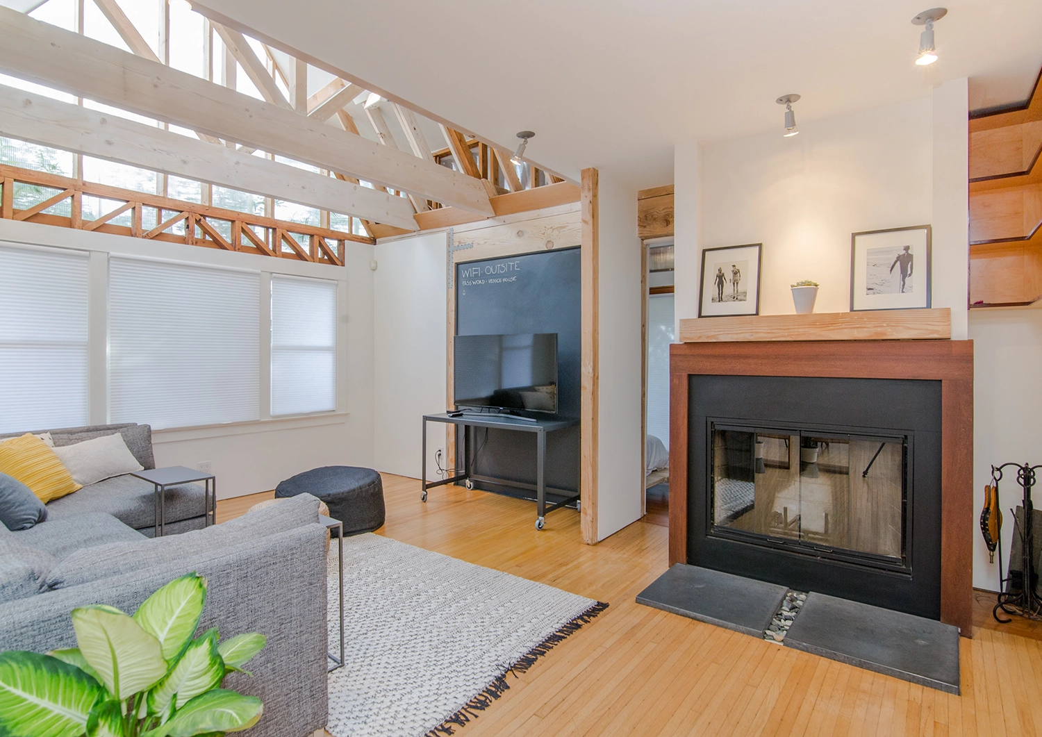 A modern living room in San Francisco with exposed wooden beams and a fireplace, remodeled by Wise Builders Remodeling, Inc., featuring a gray sofa, TV, and stylish decor.