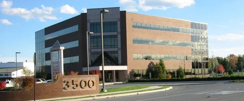 A four-story office building with large glass windows and a brick facade, surrounded by landscaping, with the address number 3500 visible in the foreground.