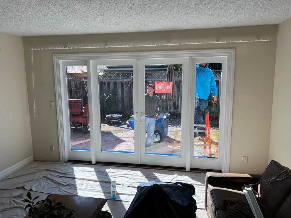 Two workers from Able Glass Company in Santa Clara, CA install a large four-panel sliding glass door from the outside of a residential home, with protective sheets covering the interior floor during the process.