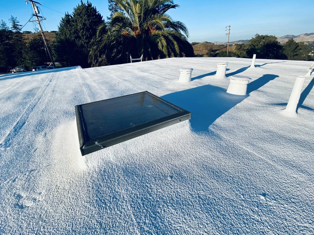 A flat white-coated roof with a skylight and several ventilation pipes under clear blue skies, showing energy-efficient roofing work by Abril Roofing, Inc. based in Martinez, CA.