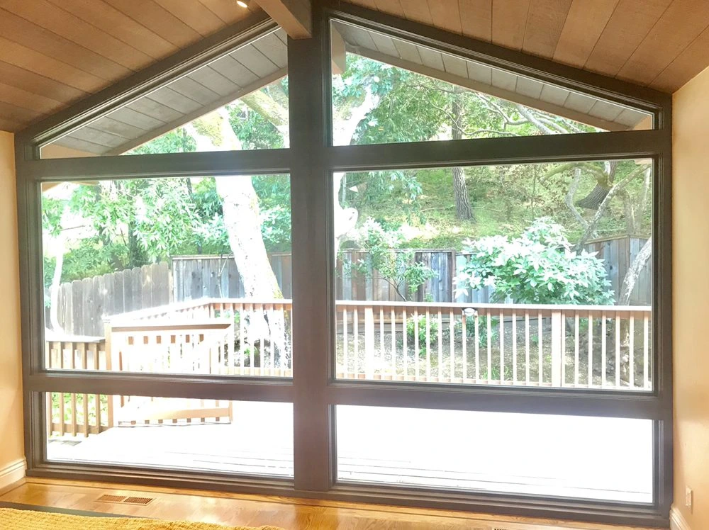 A large custom window with triangular and rectangular glass panes framed in dark metal, installed by Abstract Windows in San Carlos, CA, offering an expansive view of a deck and wooded backyard.