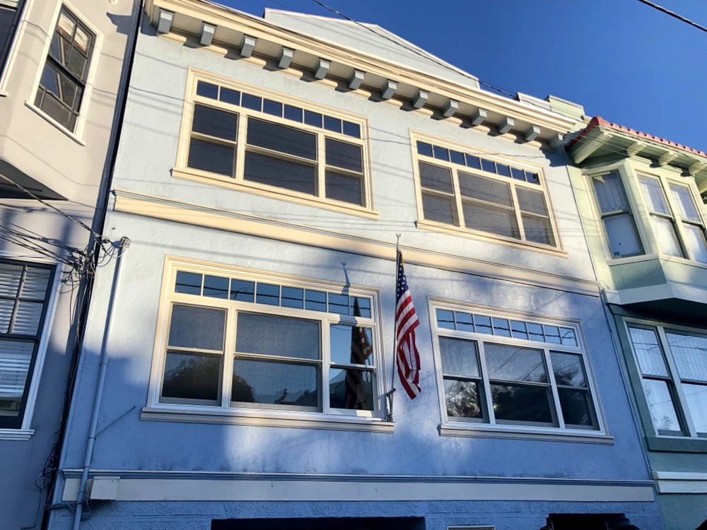 A light blue multi-story residential building with multiple symmetrically arranged cream-trimmed windows, including upper transoms, installed by Abstract Windows in San Carlos, CA, with an American flag hanging at the center.