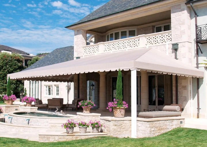 A large beige retractable awning extends over a luxurious stone patio with seating, planters, and a small fountain, installed by Acme Sunshades Enterprise Inc. in Hayward, CA.