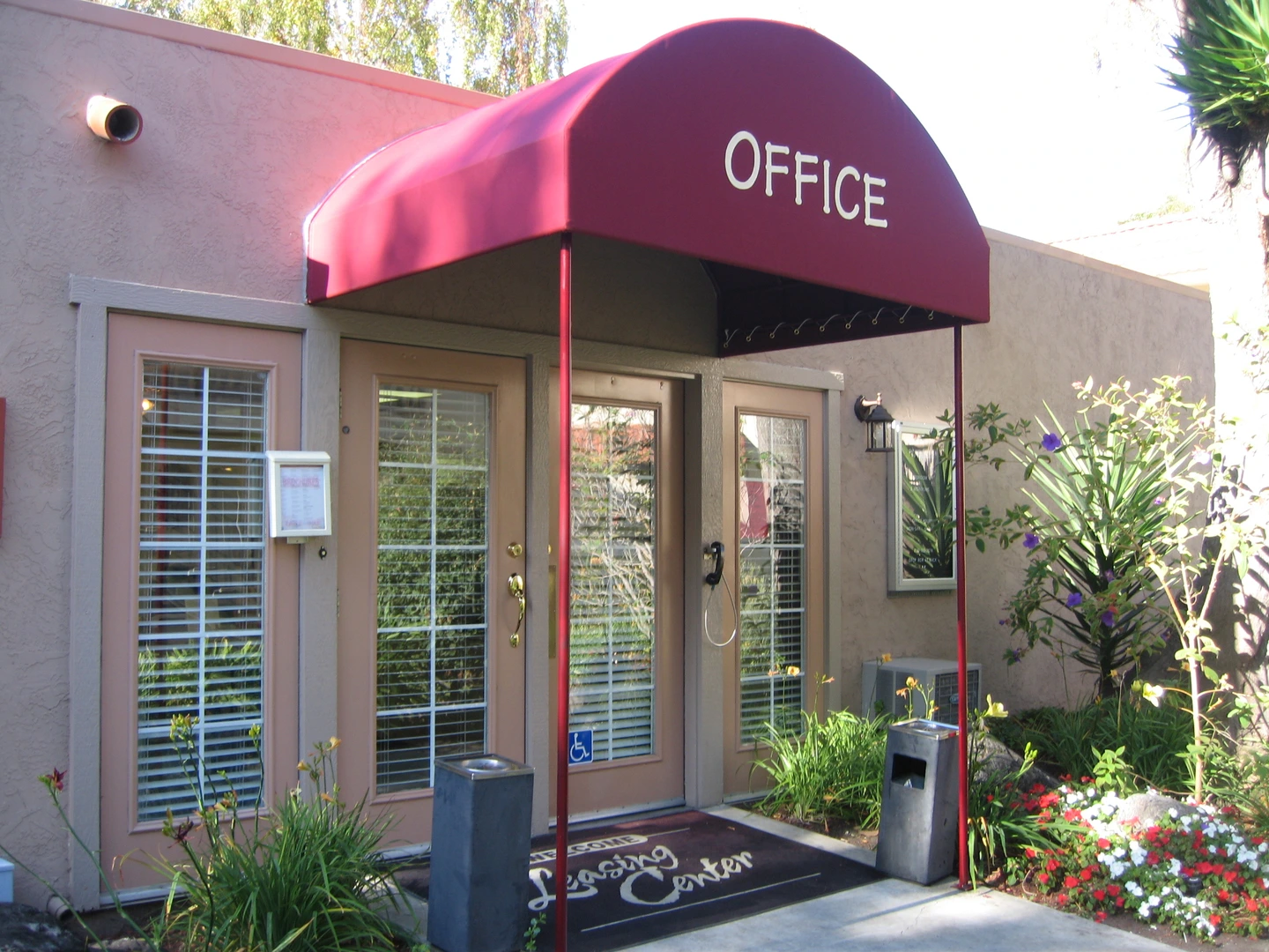 A red awning labeled “OFFICE” shades a commercial building entrance with French doors, surrounded by plants and signage, designed and installed by Acme Sunshades Enterprise Inc. in Hayward, CA.